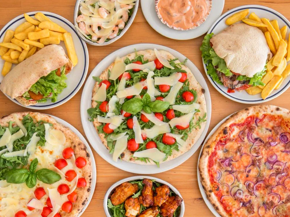 Molti piatti visti dall’alto: pizze, insalate, panini con patatine, alette di pollo e salse